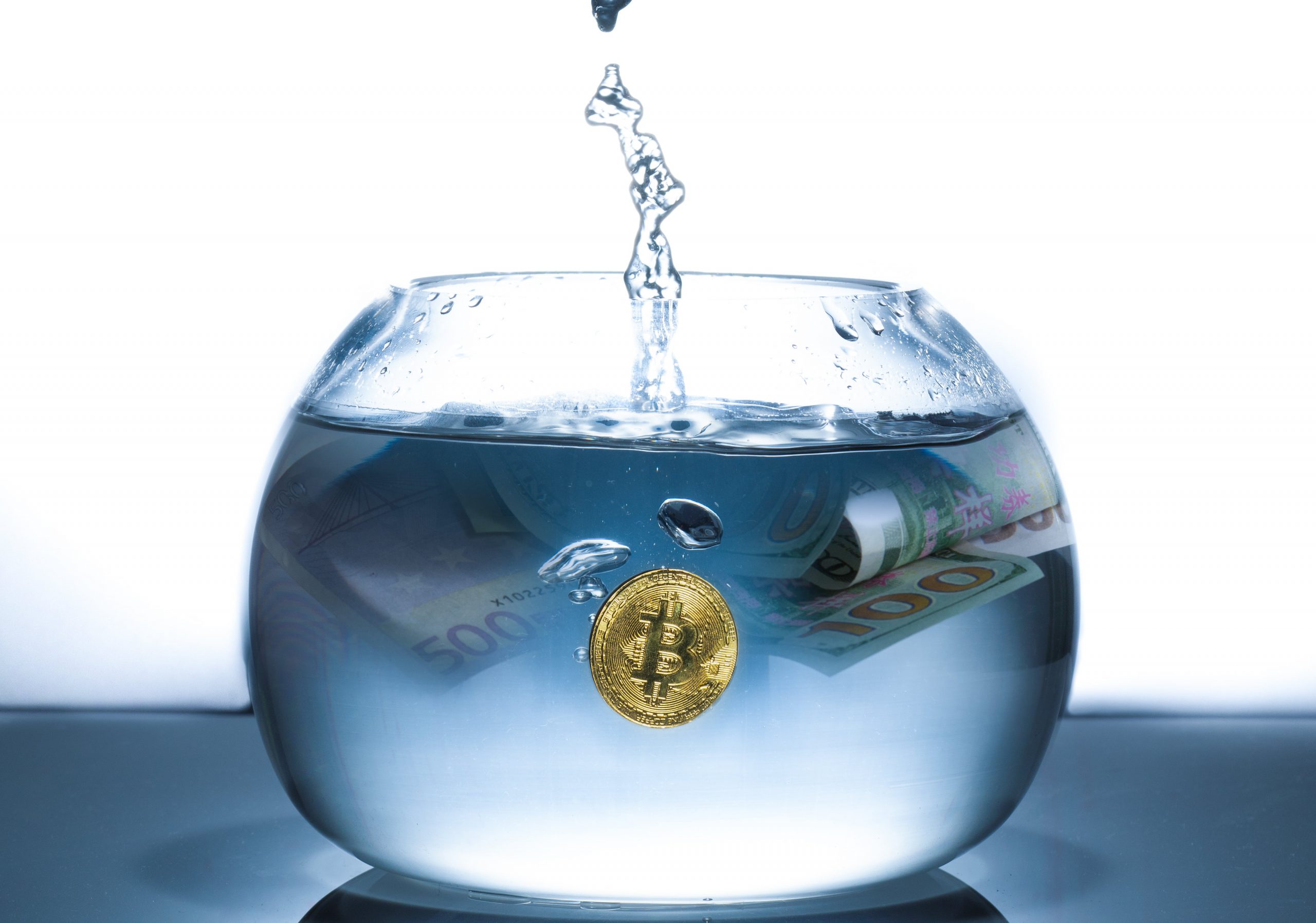 A bitcoin coin and $100 currency in a bowl being filled with water