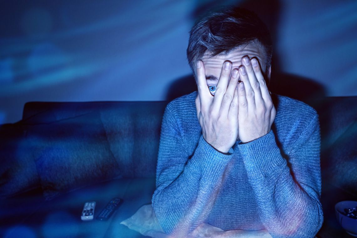 Picture of a person covering their face in fear as they watch something on the TV