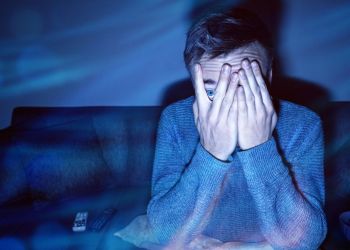 Picture of a person covering their face in fear as they watch something on the TV